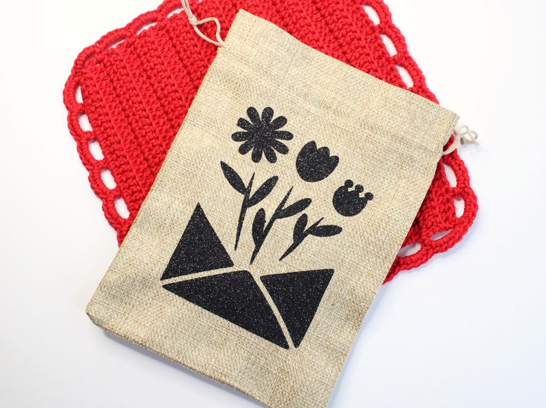 May include: A beige burlap drawstring bag featuring a black floral design, including a flower and leaves, over a geometric shape. The bag is placed on a red, crocheted, square doily with a scalloped edge.
