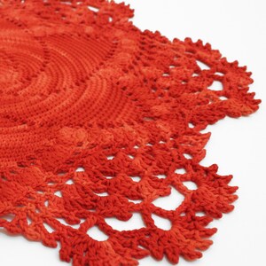 May include: A close-up of a handmade, circular, crocheted doily. The doily is a vibrant shade of orange-red, with a detailed, openwork edge. The crochet work is visible, showcasing the texture and craftsmanship of the piece. The doily is on a white background.