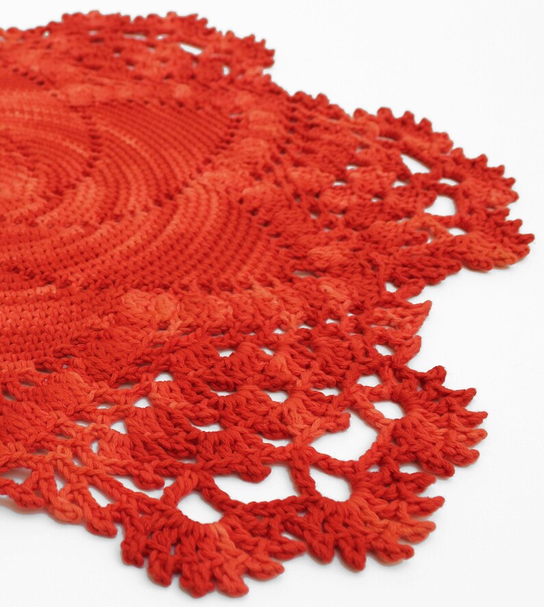 May include: A close-up of a handmade, circular, crocheted doily. The doily is a vibrant shade of orange-red, with a detailed, openwork edge. The crochet work is visible, showcasing the texture and craftsmanship of the piece. The doily is on a white background.