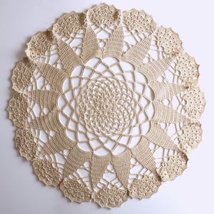 Può includere: Un centrino all'uncinetto beige di forma rotonda con un intricato motivo radiale. Il design inizia con un centro circolare denso che si trasforma in una struttura aperta a ragnatela, terminando con un bordo smerlato a forma di ventaglio.
