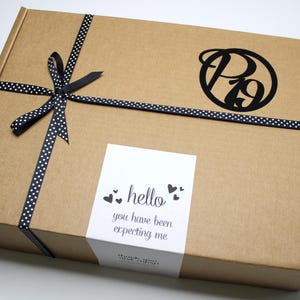 May include: A brown cardboard box tied with a black ribbon with white polka dots. The box has a black logo and a white sticker that says "hello you have been expecting me".