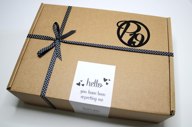 May include: A brown cardboard box tied with a black ribbon with white polka dots. The box has a black logo and a white sticker that says "hello you have been expecting me".