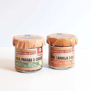 Op de afbeelding: Twee kleine glazen potjes met bruine papieren deksels. De potjes hebben etiketten met Portugese tekst. Op één potje staat "Maca, Passas & Canela" en op het andere "Figo, Laranja & Anis". De potjes staan op een wit oppervlak.