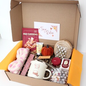 May include: A gift box filled with self-care items. Includes a mug with a bunny design, a red ginseng face mask, a small orange pumpkin, a candle, and a headband. The box also contains a card that says "Time to Relax".