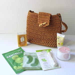 May include: A tan crochet handbag with a wooden toggle, alongside skincare items such as a face mask, eye patches, shea butter cream, and a candle. The items are displayed on a white surface.