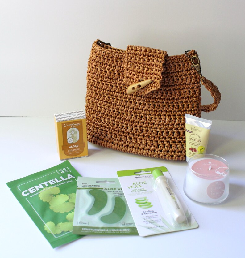 May include: A tan crochet handbag with a wooden toggle, alongside skincare items such as a face mask, eye patches, shea butter cream, and a candle. The items are displayed on a white surface.
