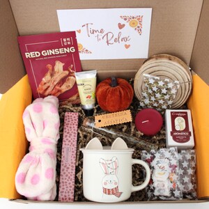 May include: A gift box filled with self-care items. Includes a red ginseng face mask, a plush pink polka-dot headband, a small pumpkin, a mug with a bunny design, a candle, and a card that says "Time to Relax".
