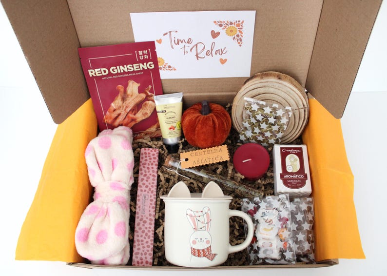 May include: A gift box filled with self-care items. Includes a red ginseng face mask, a plush pink polka-dot headband, a small pumpkin, a mug with a bunny design, a candle, and a card that says "Time to Relax".