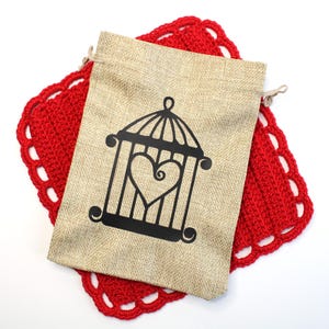 May include: A beige burlap drawstring bag with a black cage design featuring a heart shape, resting on a red crocheted doily with a scalloped edge. The bag is closed with a natural-colored drawstring.