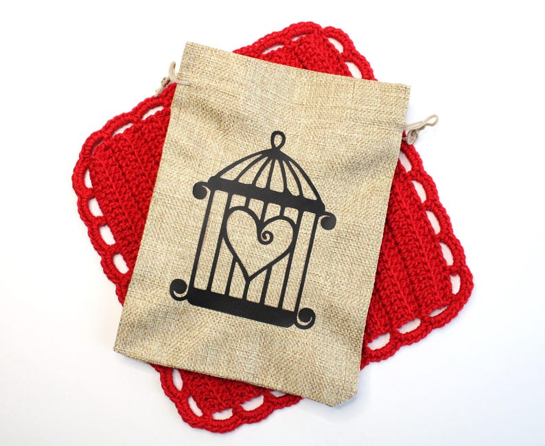 May include: A beige burlap drawstring bag with a black cage design featuring a heart shape, resting on a red crocheted doily with a scalloped edge. The bag is closed with a natural-colored drawstring.
