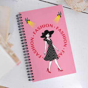 Può includere: Quaderno a spirale rosa con un design a tema moda. La copertina presenta una donna con un abito a pois e un cappello, circondata dalla parola "FASHION". Due faretti illuminano l'illustrazione. Rilegatura a spirale nera.