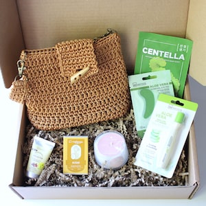 May include: A gift box containing beauty products, including a crocheted purse, face masks, a candle, and skincare items. The box contains a variety of items, including a tube of shea butter cream and a bar of soap.