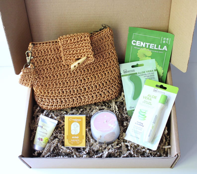 May include: A gift box containing beauty products, including a crocheted purse, face masks, a candle, and skincare items. The box contains a variety of items, including a tube of shea butter cream and a bar of soap.