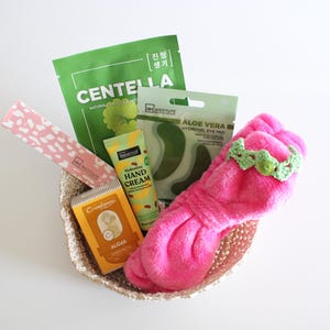 May include: A woven basket filled with beauty products. Items include a pink headband with a green crocheted band, green face masks, hand cream, and eye pads. The basket is on a white surface.