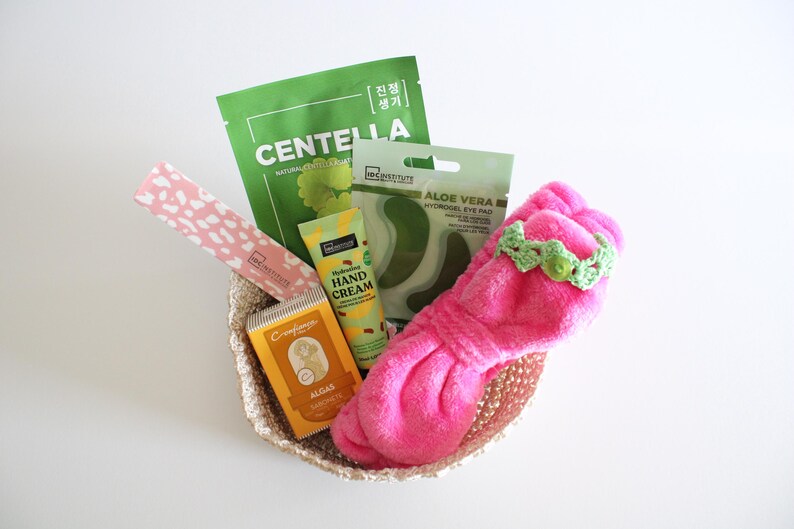 May include: A woven basket filled with beauty products. Items include a pink headband with a green crocheted band, green face masks, hand cream, and eye pads. The basket is on a white surface.