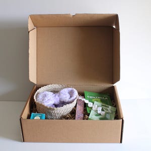Pu&ograve; includere: Una scatola di cartone aperta piena di vari oggetti. Un cestino intrecciato contiene un oggetto soffice di colore viola chiaro. Altri oggetti includono una piccola scatola blu, un pacchetto verde e un oggetto di perline bianche. La scatola &egrave; riempita con materiale di imballaggio marrone.
