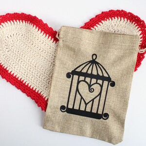 May include: A beige burlap bag with a black cage and heart design, resting on a heart-shaped, off-white crochet doily with a red border. The items are on a white background.