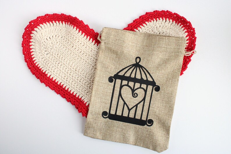 May include: A beige burlap bag with a black cage and heart design, resting on a heart-shaped, off-white crochet doily with a red border. The items are on a white background.