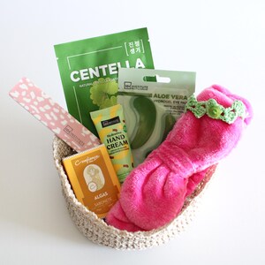 May include: A woven basket filled with beauty products. Items include a pink eye mask with a green crocheted headband, a green Centella face mask, aloe vera eye pads, hand cream, and a bar of soap.