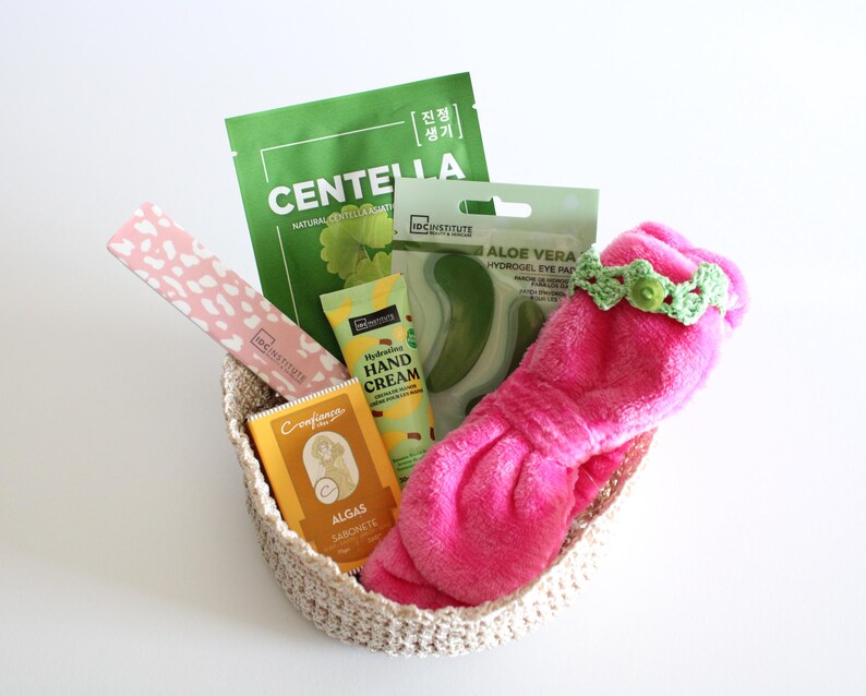 May include: A woven basket filled with beauty products. Items include a pink eye mask with a green crocheted headband, a green Centella face mask, aloe vera eye pads, hand cream, and a bar of soap.