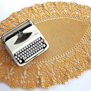 Può includere: Una macchina da scrivere portatile vintage color bianco sporco con tastiera nera e tasto di ritorno carrello rosso, posizionata sopra un grande centrino all'uncinetto ovale color giallo senape con un intricato motivo a foglie.