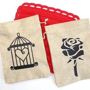 May include: Two beige burlap drawstring bags with black graphic designs. One bag features a cage with a heart inside, and the other has a rose. A red crocheted square is behind the bags.