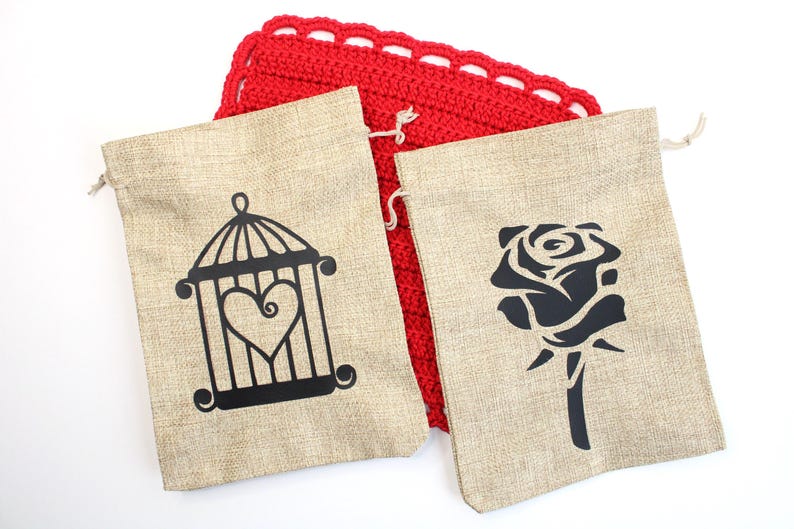 May include: Two beige burlap drawstring bags with black graphic designs. One bag features a cage with a heart inside, and the other has a rose. A red crocheted square is behind the bags.