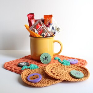 May include: A yellow mug filled with packets of coffee and candy, sitting on a crocheted orange coaster. Decorative Halloween-themed coasters with spider and pumpkin designs are also present, along with small green and purple embellishments.