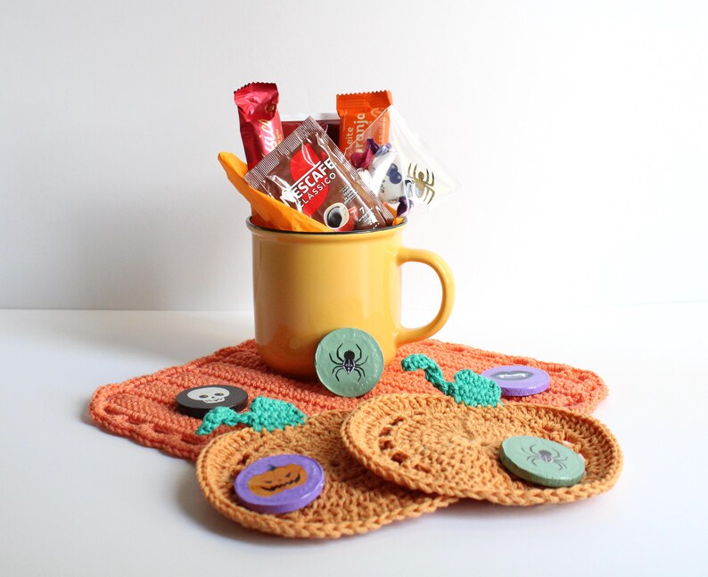 May include: A yellow mug filled with packets of coffee and candy, sitting on a crocheted orange coaster. Decorative Halloween-themed coasters with spider and pumpkin designs are also present, along with small green and purple embellishments.