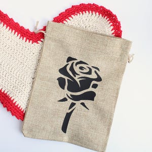 May include: A beige burlap bag with a black rose design, next to a white and red crocheted heart-shaped doily. The bag has a drawstring closure. The heart-shaped doily has a red border.
