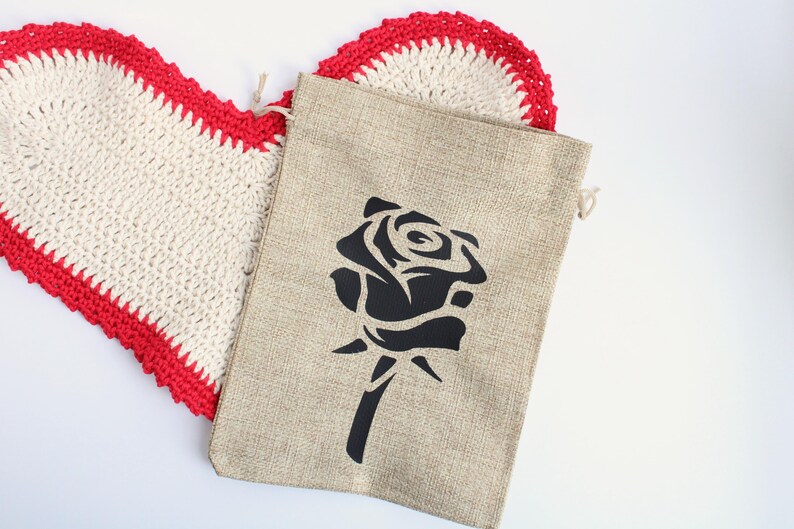 May include: A beige burlap bag with a black rose design, next to a white and red crocheted heart-shaped doily. The bag has a drawstring closure. The heart-shaped doily has a red border.