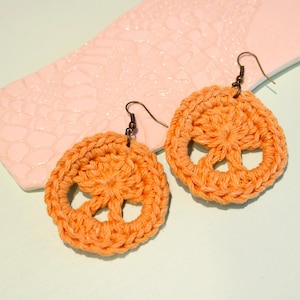 May include: A pair of orange crochet earrings in a peace sign design. The earrings are made of a thick, textured yarn and have bronze-colored ear wires. The earrings are displayed on a light pink, textured ceramic dish against a light green background.