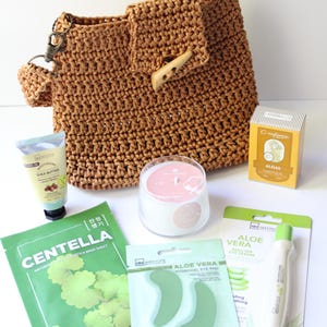 May include: A brown crocheted handbag with a wooden button closure, surrounded by skincare products. These include shea butter cream, a Centella face mask, aloe vera eye pads, a roll-on eye serum, a candle, and algas soap.