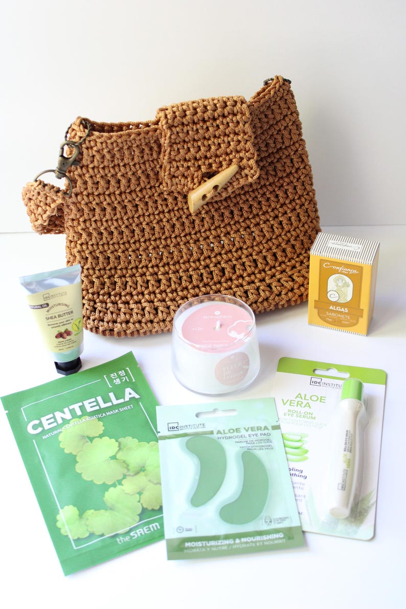 May include: A brown crocheted handbag with a wooden button closure, surrounded by skincare products. These include shea butter cream, a Centella face mask, aloe vera eye pads, a roll-on eye serum, a candle, and algas soap.