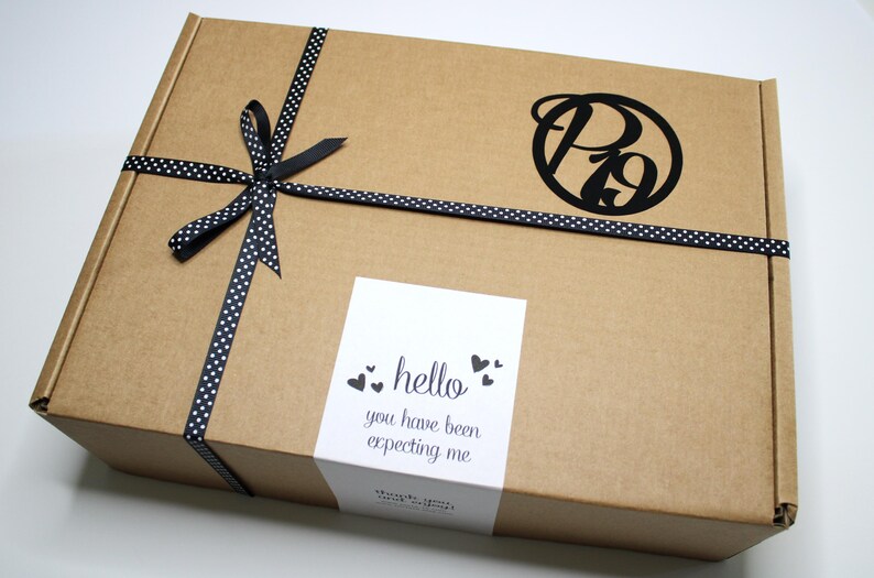 May include: A brown cardboard box tied with a black and white polka dot ribbon. The box features a black logo and a white sticker with the text "hello you have been expecting me".