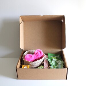 May include: A brown cardboard box with an open lid, containing a woven basket with a pink fluffy bow, green packets, and other small items. The box is on a white surface.