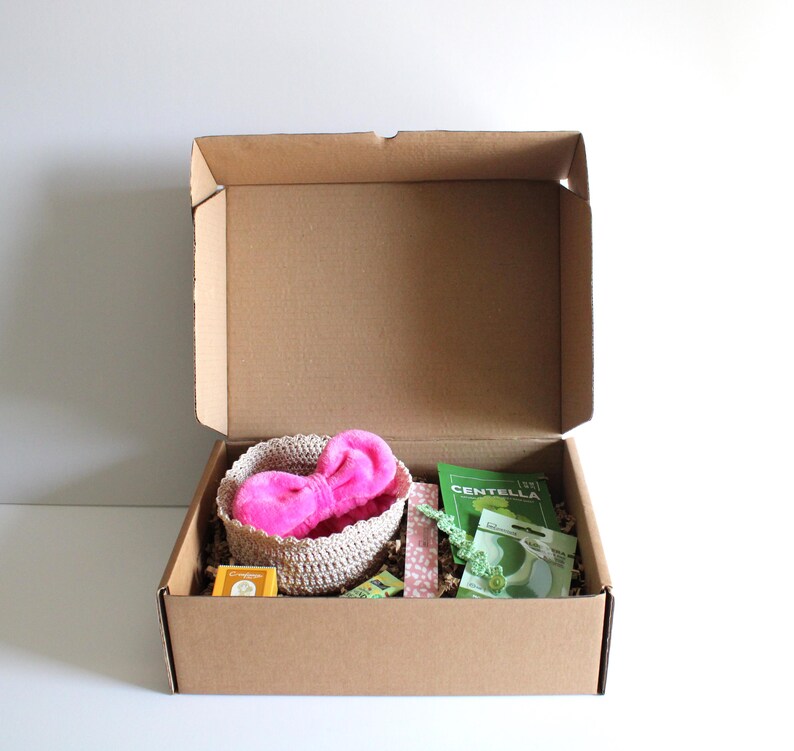 May include: A brown cardboard box with an open lid, containing a woven basket with a pink fluffy bow, green packets, and other small items. The box is on a white surface.