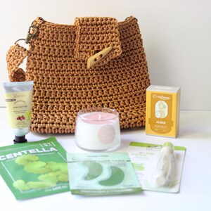 May include: A brown crocheted shoulder bag with a wooden button closure, alongside skincare items such as shea butter cream, eye patches, and a candle. A box of algae soap is also visible.