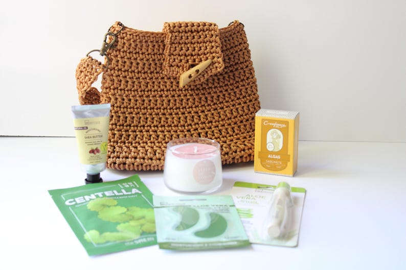 May include: A brown crocheted shoulder bag with a wooden button closure, alongside skincare items such as shea butter cream, eye patches, and a candle. A box of algae soap is also visible.