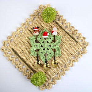 May include: A festive Christmas-themed arrangement featuring three decorative spoons with reindeer and snowman toppers. The spoons rest on a green crocheted snowflake, set atop a beige crocheted doily, with green pom-poms and decorative sticks.