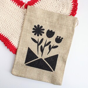 May include: A beige burlap gift bag with a black floral design. The design features a flower and leaves emerging from an envelope. A crocheted heart with a red border is in the background.