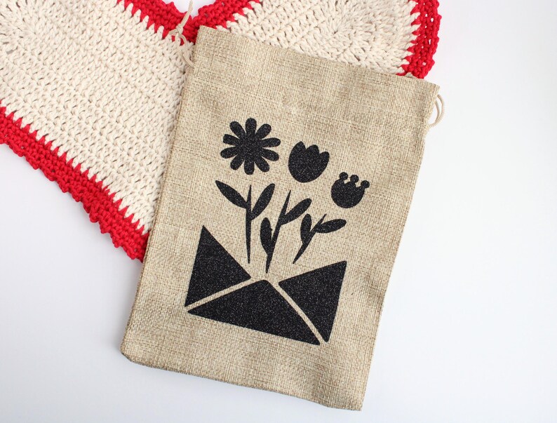 May include: A beige burlap gift bag with a black floral design. The design features a flower and leaves emerging from an envelope. A crocheted heart with a red border is in the background.