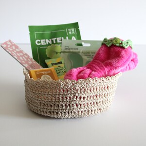May include: A beige crochet basket filled with beauty products. Items include a pink fuzzy sock with a green floral trim, a pink nail file, and green and yellow packaged skincare items. The basket is on a white surface.