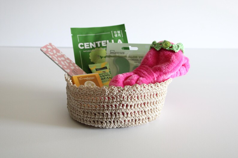 May include: A beige crochet basket filled with beauty products. Items include a pink fuzzy sock with a green floral trim, a pink nail file, and green and yellow packaged skincare items. The basket is on a white surface.