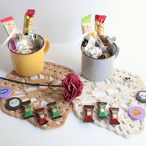 May include: Two mugs filled with candy and treats, presented on crocheted doilies. One mug is yellow, the other gray. Halloween-themed pins and a dark red flower add decorative touches. Candy bars and wrapped sweets are visible.