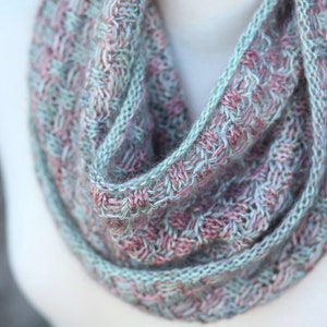 May include: A close-up of a knitted scarf in shades of pink and green. The scarf is made with a textured stitch pattern and is folded in a loop.