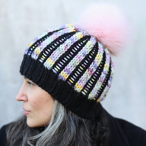 Könnte beinhalten: Eine schwarze Strickmütze mit mehrfarbigen Streifen und einem rosa Kunstfellbommel.