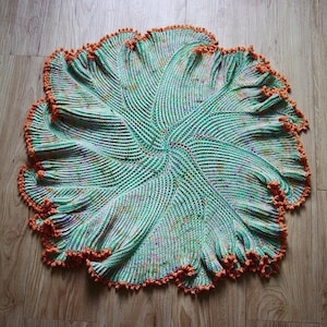 May include: A crocheted baby blanket in a green and orange color scheme. The blanket is circular and has a scalloped edge.