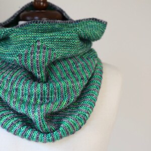May include: A green and gray knitted cowl scarf with a hood. The scarf is made of a soft, textured yarn and features a ribbed design. The hood is folded over, and the scarf is displayed on a mannequin.