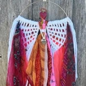 Puede incluir: Un colgante de pared hecho a mano con un marco circular de metal dorado y un diseño de ángel. El ángel tiene alas de ganchillo blancas, detalles de tela colorida y una maceta de terracota con vegetación. El colgante se suspende con un cordón.